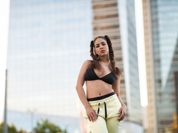 Young self assured slim ethnic sportswoman in bra and pants standing looking at camera in city park with hands in pockets against modern skyscrapers