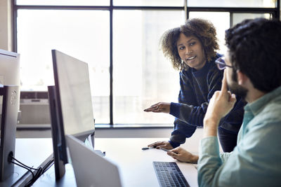 Colleagues discussing over desktop computer on desk against window in office