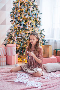 Full length of girl sitting against christmas tree at home