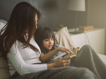 Smiling mother and girl reading book while sitting on bed at home