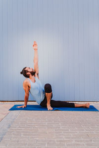 Full length of man performing yoga on exercising mat against wall