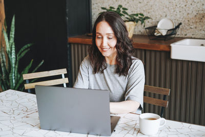 Adult smiling brunette woman having breakfast with opened laptop in kitchen at the home