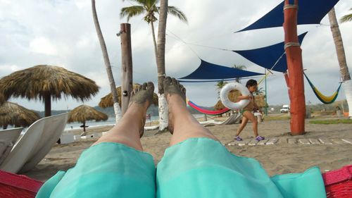 Low section of woman lying in hammock while boy walking with inflatable ring at beach