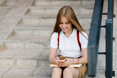 A cheerful teenage girl sits on the steps of the school, writes a message on her phone and wait