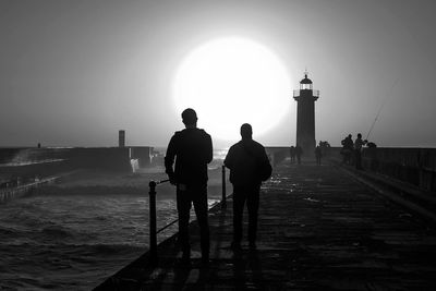 Silhouette people at pier leading towards lighthouse on sunny day