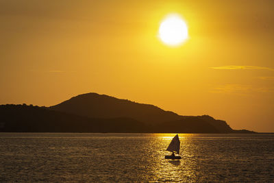 Silhouette people on sailboat by sea against orange sky