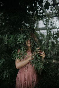 Female standing in lush greenhouse and hiding face behind branch of tree with green foliage