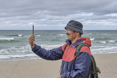 Man standing at beach and shooting video on phone against sea and sky