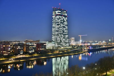 Illuminated buildings by river against blue sky at night