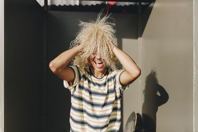 Playful woman with hands in curly blond hair amidst metallic walls on sunny day