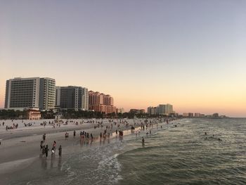 People on beach by buildings against sky during sunset