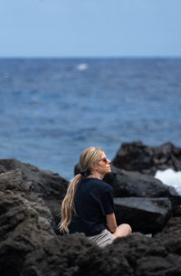 Rear view of woman sitting on rock by sea against sky