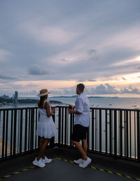 Rear view of woman standing by railing against sea during sunset