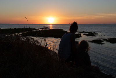 Woman and her daughter sitting on shore at beach against sky during sunset