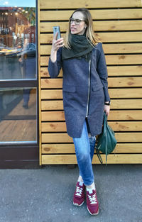 Young woman in coat and jeans indignantly looks at smartphone screen in the street near wooden wall