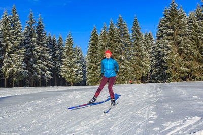 Full length of woman looking away while skiing on snow covered land against trees and sky