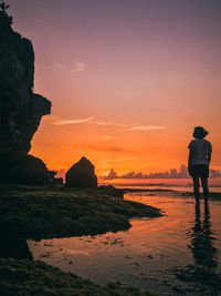 Rear view of silhouette man on rock at beach against sky during sunset