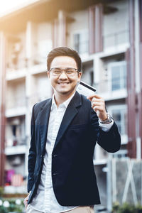 Portrait of smiling businessman holding credit card against building