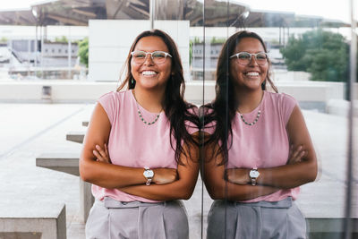 Delighted young ethnic woman with long dark hair in stylish clothes and eyeglasses smiling brightly and looking away while leaning on glass mirrored wall with crossed arms on street