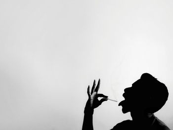 Side view of silhouette man holding lit cigarette by tongue against white background