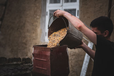 A young guy pours wheat and corn from a bucket into a crusher.