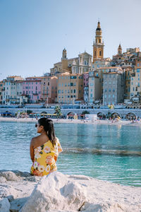 Woman sitting by buildings in city against sky