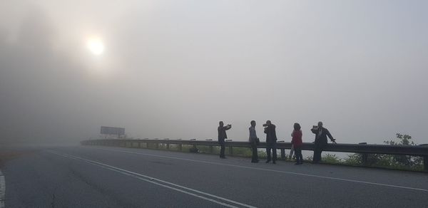 People on road against crash barrier and sky during foggy weather