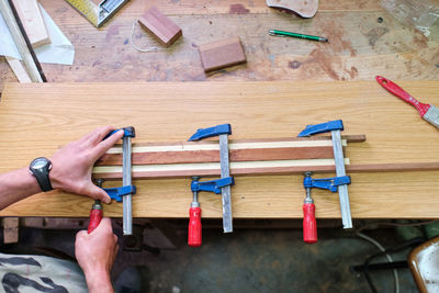 Overhead view of crop unrecognizable male carpenter fixing wooden sticks glued together with clamps at workbench with professional tools