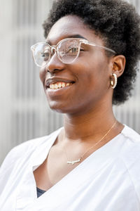 Delighted african american female in white wear and glasses and work uniform looking away with smile while standing on street near wall