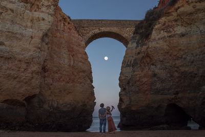 Back view of unrecognizable male traveler embracing happy girlfriend on while relaxing together on famous praia de estudantes sandy beach and admiring ocean against cloudless evening sky