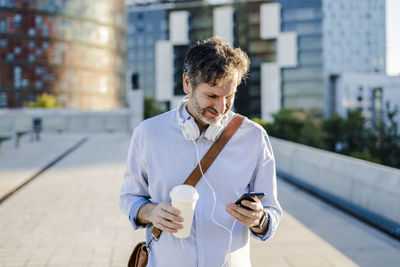 Mature man with takeaway coffee and headphones using cell phone in the city