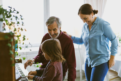 Happy mother and great grandfather looking at boy playing piano in house