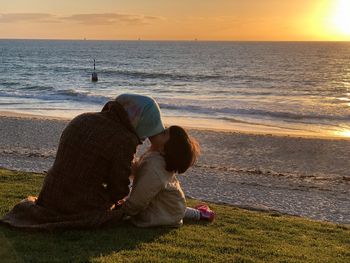 Rear view of woman sitting with daughter at beach during sunset
