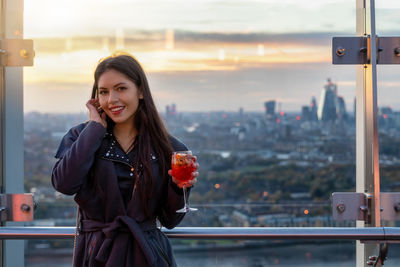 Portrait of smiling young woman holding drink while standing against window during sunset
