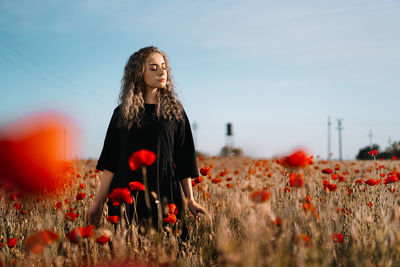 Beautiful young woman standing by poppy flowers on field against sky