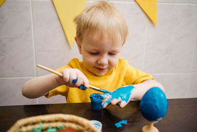 Little boy draws blue paint on his hand, paints an easter egg