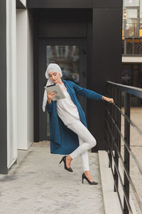 Full length portrait of woman standing on footpath against building