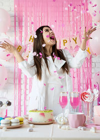 Woman in white party clothes preparing birthday table throwing pink confetti