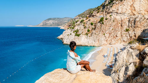 Rear view of woman sitting on rock by sea against clear blue sky