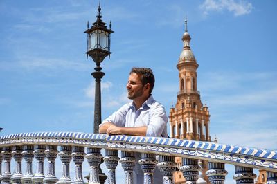 Man leaning on railing with historic tower in background against blue sky
