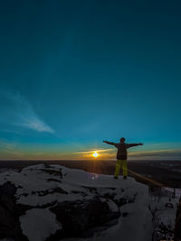 Snowboarder stands on end of mountain, looks into the distance at sunset