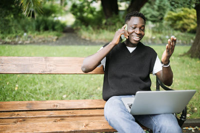 Young african man talking on phone, working on laptop in park, happy, pleased