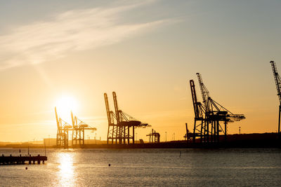 Silhouette cranes at commercial dock against sky during sunset