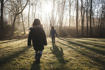 Rear view of sisters walking on grassy field in forest during sunny day