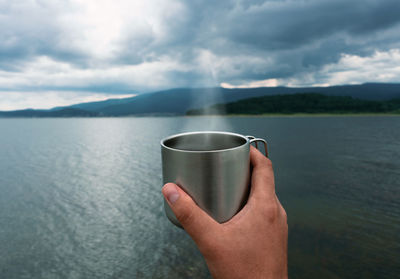 Midsection of person holding drinking water against lake