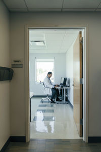 Side view of doctor using desktop computer while working in hospital seen through doorway
