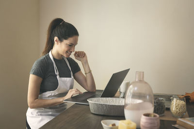 Smiling woman reading recipe on laptop while preparing food at table