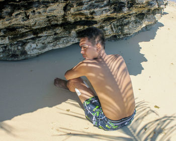 Full length of shirtless man sitting at beach with shadow of palm tree on back