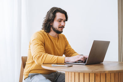 Young bearded brunette business man in casual yellow longsleeve working on laptop in modern office