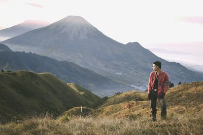 Young man standing on field while looking at mountains against sky